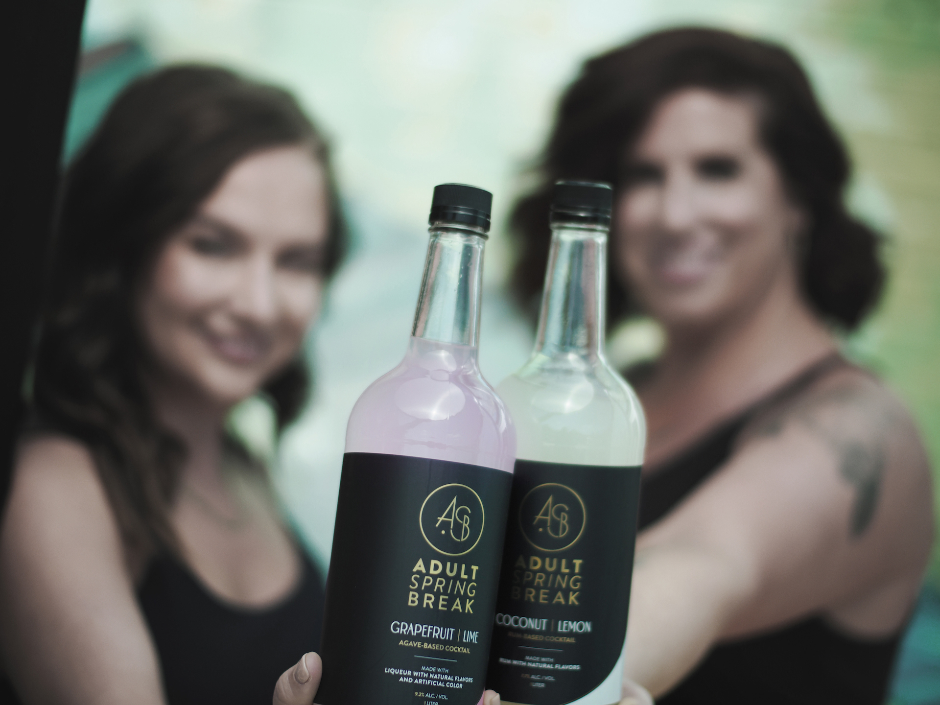 Founders Kendall and Casey out of focus holding two Adult Spring Break bottles in focus. 